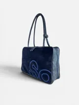 Bolso de mano tipo maletín, confeccionado en cuero natural y gamuza negra con bordado decorativo en tono azul. Cuenta con doble asa superior, cierre principal con doble cursor y base con topes metálicos.