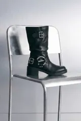 Bota media de cuero vegano color negro, con taco cuadrado y puntera redondeada. Cuenta con detalles de hebillas metálicas y correas decorativas en la caña y alrededor del tobillo.
