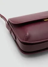 Bolso tipo bandolera de tamaño pequeño con acabado efecto piel en color bordó. Presenta una solapa frontal con cierre metálico a presión, costuras visibles en contraste y una correa larga ajustable con herrajes dorados.