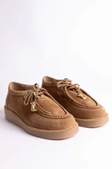 Zapato tipo mocasín de gamuza color tostado, con suela de plataforma plana y cordones. Presenta un detalle decorativo de dije metálico dorado en forma de bota en cada zapato.