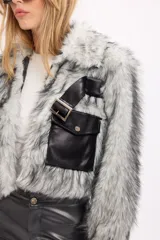 Campera de pelo sintético en tonos gris y blanco, con diseño de corte corto. Presenta un bolsillo frontal tipo cargo en cuero sintético negro con solapa y una correa decorativa con hebilla metálica sobre el pecho.