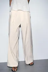 Pantalón de tiro alto en color beige, con diseño de pierna ancha y pinzas frontales. Cuenta con bolsillos laterales, cierre frontal con cremallera y gancho metálico, y un lazo ajustable en la cintura del mismo tejido.