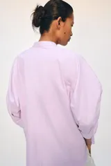 Camisa confeccionada en hilatura de algodón de color rosado claro, con cuello mao y cierre frontal parcial con botones a contraste. Presenta mangas por debajo del codo abullonadas y bajo asimétrico con aberturas laterales.