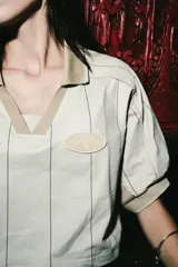 Camiseta crop de manga corta con cuello polo, confeccionada en tela técnica tipo dry fit. Presenta un diseño a rayas verticales finas en color beige, con paneles laterales de malla en tono camel y detalles de parches bordados en el frente.