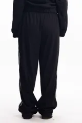 Pantalón deportivo negro de calce relajado con cintura elástica. Presenta una franja lateral gruesa de color gris claro con bordes blancos. Se observa un pequeño bordado en la zona del bolsillo con la palabra "Delosalto".