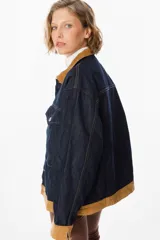 Chaqueta de denim azul oscuro con cuello y puños en pana color tostado. Presenta cierre frontal con botones metálicos y dos bolsillos frontales con solapa.