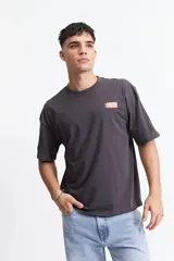 Remera de manga corta color gris oscuro, con corte holgado, hombros caídos y escote redondo. Presenta un logo estampado en el pecho con la inscripción "GALLERY DEPT." en color naranja.