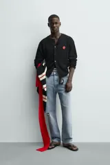 Cárdigan de punto negro, corte relajado, con cuello redondo, manga larga y cierre frontal de botones oscuros. Presenta un parche bordado en forma de dado rojo con corazones blancos en el lado izquierdo del pecho.