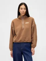 Buzo de polar tipo sherpa color marrón, con cuello alto y cierre frontal parcial. Presenta el logo de la marca bordado en el pecho y detalles de costuras reforzadas en hombros y mangas.