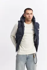 Chaleco puffer acolchado color azul marino, con capucha fija, cierre frontal con solapa de botones a presión y bolsillos laterales con cierre. Presenta un parche con el logo de la marca en el pecho.