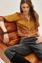 Chaqueta corta color mostaza con mangas abullonadas y bordados étnicos en tonos rosados y rojos en el frente, puños y hombros. Presenta un cierre frontal con detalles de cuentas.
