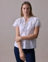 Blusa blanca de algodón marca Cupio, con cuello a la base, escote en V y lazo de ajuste. Presenta bordado en la parte delantera y mangas cortas amplias con aplique de puntilla.