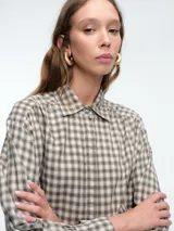 Camisa de corte clásico con estampado de cuadros vichy en tonos azul y blanco. Presenta cuello camisero, cierre frontal con botones ocultos y mangas largas con puños abotonados. Incluye un detalle de broche metálico dorado en el cuello.