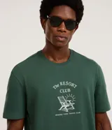 Remera de algodón color verde oscuro, con cuello redondo y manga corta. Presenta una estampa frontal en color blanco con el texto 'The Resort Club' y una ilustración de una silla de playa y el sol.