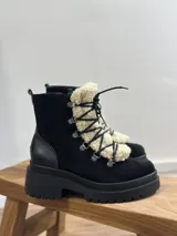 Bota tipo borcego de cuero marrón con suela track negra de goma. Presenta un aplique frontal desmontable de corderito sintético color crema y cordones negros con ganchos metálicos.