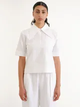 Camisa blanca de manga corta con cuello camisero grande y marcado, cierre frontal oculto y bolsillo ojal en el pecho.