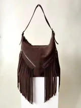 Bolso tipo hobo color tostado, hecho de cuero con flecos largos en el frente y dos bolsillos con cierre. Tiene una manija regulable con hebillas metálicas.