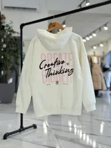 Canguro oversize de color beige claro con capucha, mangas largas y puños elastizados. Presenta un diseño gráfico en la espalda con el texto 'Creative Thinking' en tipografía cursiva negra sobre un fondo de letras grandes en tono rosado.