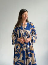 Vestido largo azul con estampado de hojas en tonos beige y blanco, escote en V con botones frontales, mangas largas amplias y diseño escalonado.