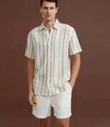 Camisa de hombre confeccionada en 100% algodón con textura, de manga corta y cuello inglés. Presenta un diseño de rayas verticales en tonos beige, blanco y marrón, con cierre frontal de botones.