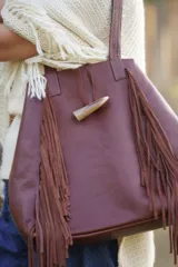 Bolso tipo tote de cuero color marrón, con diseño de flecos laterales y cierre frontal mediante un botón de guampa. Cuenta con doble asa de hombro.