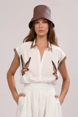 Camisa blanca de poplin de algodón con cuello camisero, abotonadura frontal y mangas cortas. Presenta un estampado de aves rapaces en el frente y galones decorativos en tonos marrones y beige en cuello y mangas.