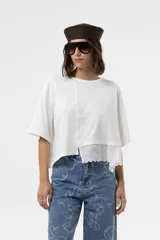 Remera blanca de calce relajado y silueta amplia, con mangas cortas, hombros caídos, corte levemente cropped y detalle de broderie en la parte frontal inferior.