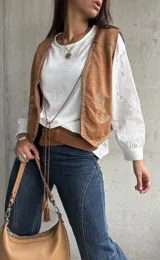 Chaleco de gamuza color camel con bordados decorativos en hilo claro y cierre frontal mediante lazos con borlas. Presenta un diseño de inspiración boho con terminaciones delicadas.