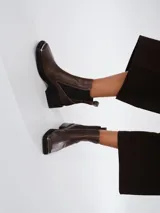 Botas tipo chelsea de cuero color marrón oscuro con acabado brillante. Presentan punta cuadrada con detalle de puntera metálica, elásticos laterales y taco cuadrado bajo.