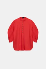 Camisa de algodón con cuello redondo y cierre frontal de botones a contraste. Presenta mangas abullonadas por debajo del codo y bajo asimétrico con aberturas laterales.