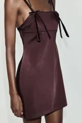 Vestido mini confeccionado en tejido satinado color bordó, de escote recto con tirantes finos y detalle de lazos aplicados en el delantero.