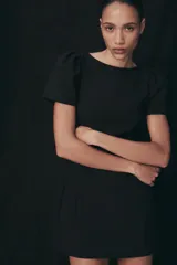 Vestido corto de color negro, con cuello redondo, manga corta abullonada y cierre de cremallera oculta en la espalda.