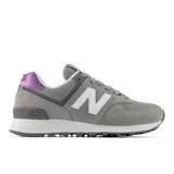Championes urbanos New Balance modelo 574, color gris con detalles en blanco y panel metalizado violeta en el talón. Confeccionados en gamuza y malla, cuentan con entresuela ENCAP para mayor amortiguación y suela de goma.