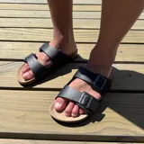 Sandalia tipo Birkenstock con doble tira ajustable de cuero sintético negro y hebillas metálicas. Cuenta con plantilla anatómica de corcho y suela de goma.