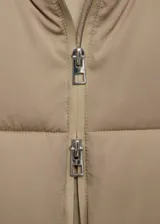 Campera puffer acolchada de color beige, con diseño recto, cuello alto y capucha integrada. Cuenta con cierre frontal de cremallera doble, mangas largas con puños elásticos y dos bolsillos laterales con cierre. Acabado repelente al agua.
