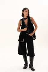 Chaleco negro de gamuza con diseño sin mangas, decorado con ojales metálicos plateados a lo largo de los bordes frontales y sisas.