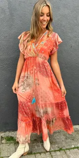 Vestido largo anaranjado con estampado estilo paisley y geométrico, escote en V con bordados multicolor, mangas cortas con volados, cordón ajustable en la cintura con borlas turquesas y falda escalonada.