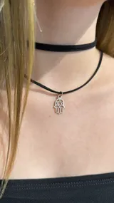 Gargantilla tipo choker compuesta por una cinta negra de tela y un dije metálico plateado con forma de mano de Hamsa que incluye una estrella de David en su interior.