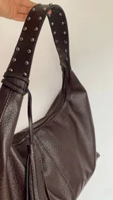 Cartera estilo hobo color chocolate, confeccionada en eco cuero con textura granulada. Presenta una correa ancha fija con detalles de tachas metálicas y un colgante de borla lateral. Incluye cierre superior y herrajes plateados.