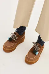 Zapato de plataforma estilo mocasín, confeccionado en cuero marrón con detalles de gamuza en el talón y la lengüeta. Presenta cordones estampados, flecos decorativos y pompones en tonos verde y celeste.