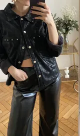 Camisa negra con tachas plateadas en cuello y solapas, cierre con botones metálicos y mangas largas con puños abotonados.