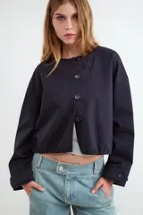 Campera corta oversize de color negro, confeccionada en algodón, con mangas largas, cuello redondo, cierre frontal de botones y bajo con elástico fruncido.