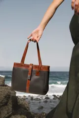 Cartera tipo tote de cuero vacuno, con diseño bicolor en gris y marrón. Cuenta con doble compartimento, cierre metálico, asas largas y una correa central decorativa con cierre de broche metálico. Incluye un detalle de etiqueta colgante de cuero.