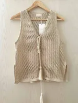 Chaleco tejido en punto calado color beige con destellos de lurex. Presenta escote profundo en V con cordones frontales para atar terminados en borlas, corte recto y aberturas laterales en el ruedo.
