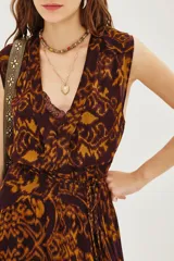 Vestido largo sin mangas con escote en V profundo y lazo ajustable en la cintura. Presenta un estampado geométrico en tonos marrón y ocre sobre fondo oscuro, con falda amplia y fluida.
