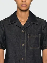 Vestido corto de denim oscuro con cuello camisero, cierre frontal de botones dorados y bolsillos frontales tipo parche con solapa. Presenta costuras en contraste y mangas cortas.