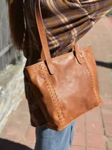 Cartera tipo tote de cuero color camel, con diseño estructurado, costuras decorativas laterales y doble asa superior para el hombro.