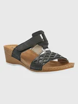 Sandalia de plataforma baja con diseño de tiras negras. La tira frontal presenta un patrón geométrico calado con pequeñas tachas metálicas, conectada a una tira superior ajustable mediante velcro a través de una argolla metálica. La plantilla es de color marrón y la suela tiene efecto madera.
