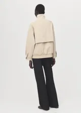 Chaqueta oversize de algodón color beige, con cuello alto, cierre frontal oculto con cremallera, mangas largas con puños abotonados, bolsillos laterales y bajo fruncido elástico.