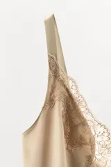 Vestido mini de tejido satinado color beige, con escote en pico adornado con encaje, tirantes anchos y detalle de encaje bordado en la cintura. El bajo presenta un acabado con volantes.